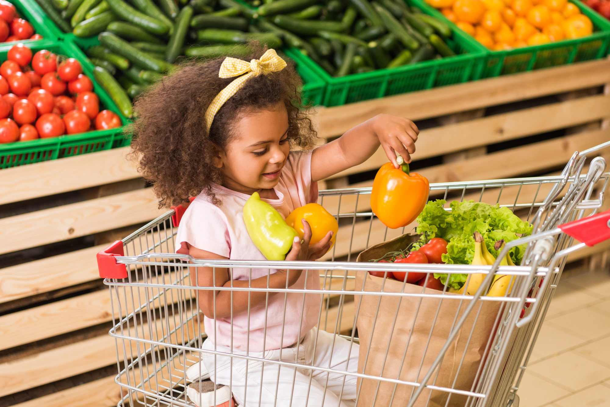 Smiling girl putting bell peppers in paper bag while sitting in shopping trolley in supermarket