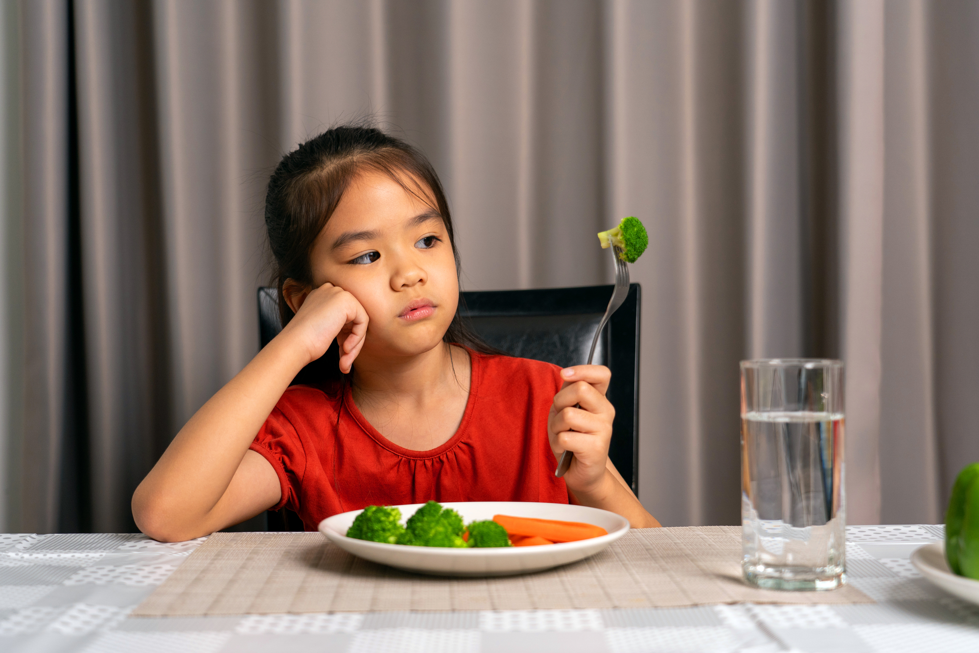 a child sitting at a table looking at a piece of broccoli on their fork with distaste 
