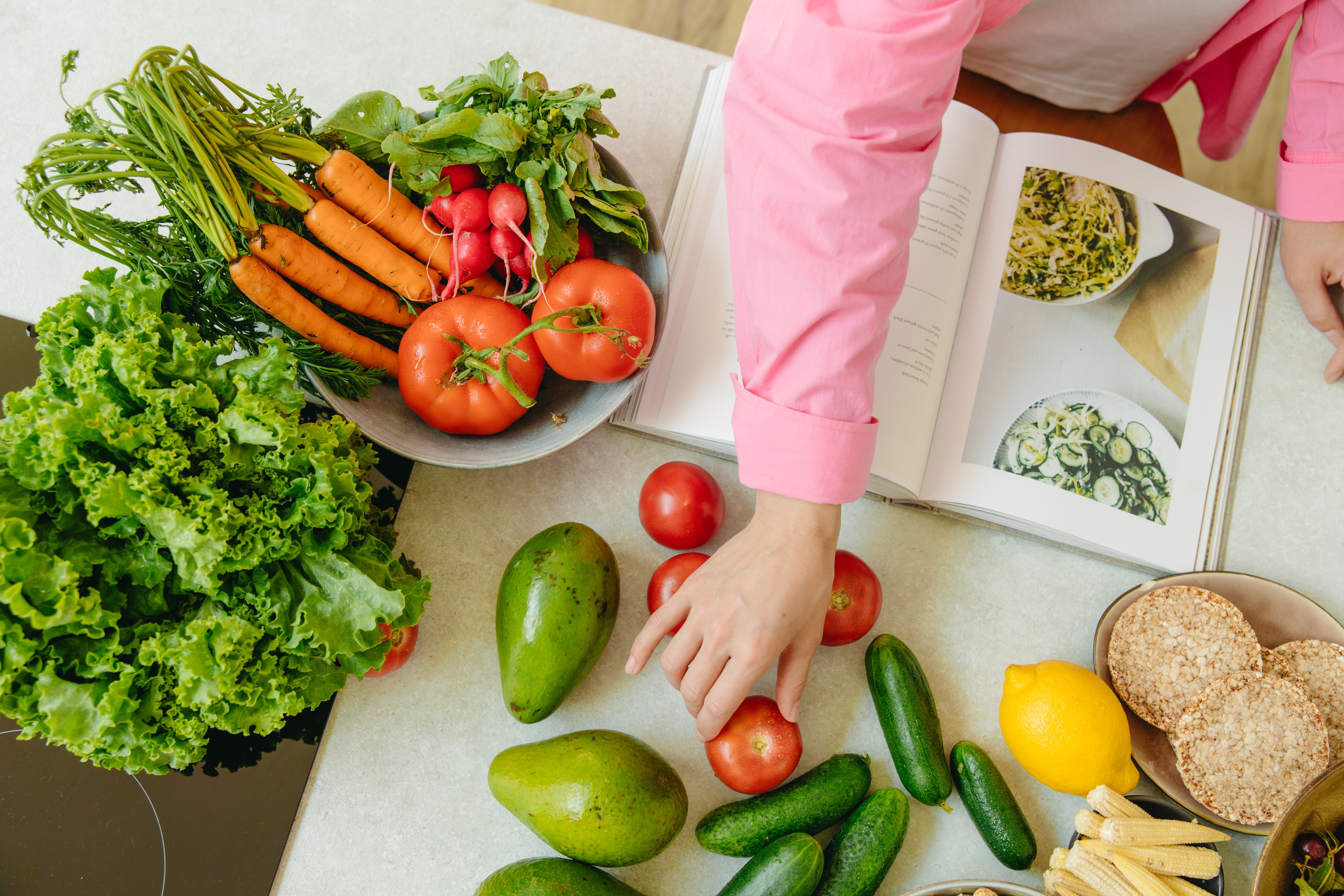 Kitchen counter with fruits, vegetables and a cookbook. There is also an outstretched hand reaching over the counter 