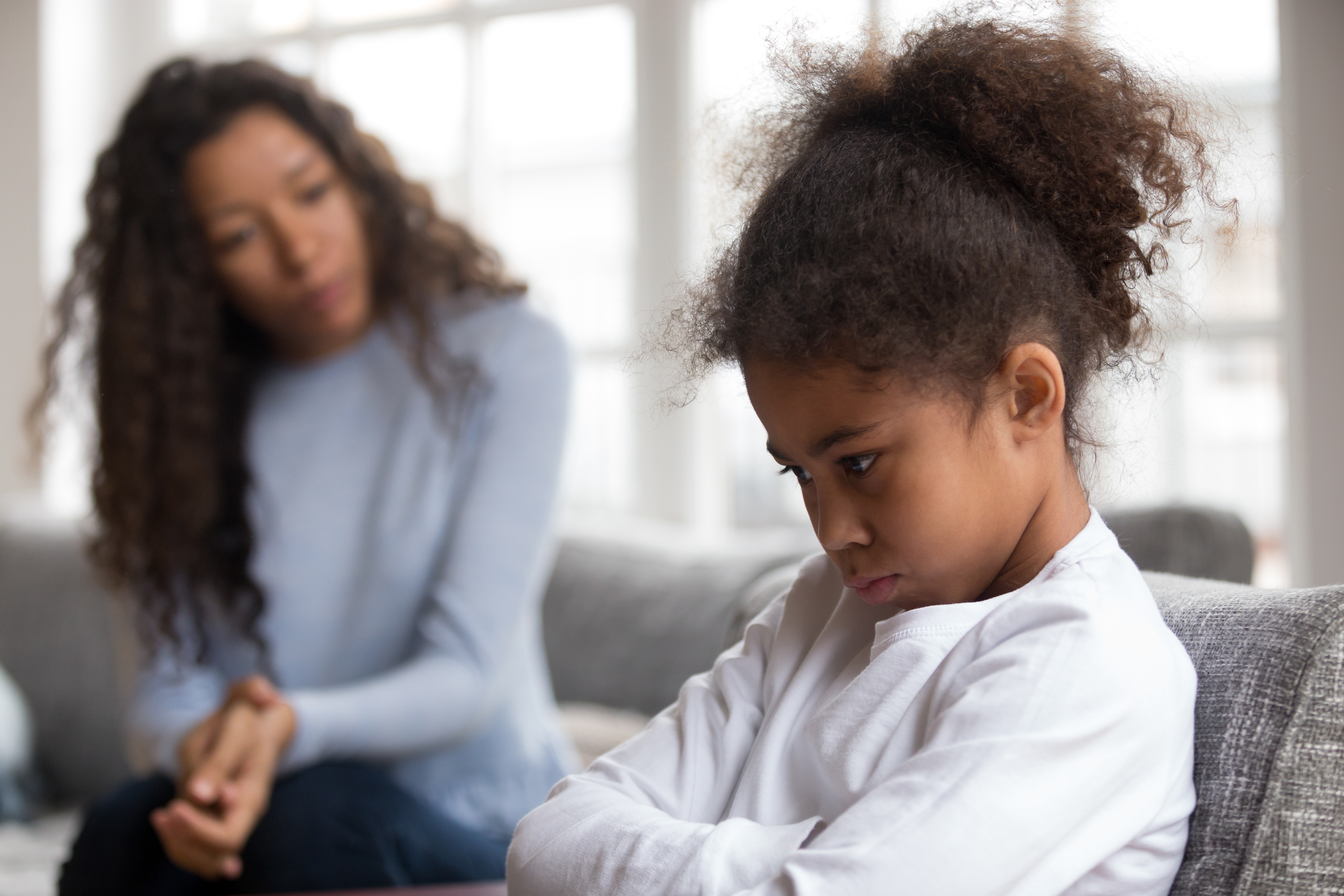 Child mental health: Little girl frowning with arms crossed, refusing to talk to mom in background