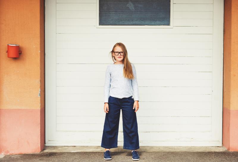 pre-adolescent girl standing in front of garage door wearing blue top, denim culottes and eyeglasses