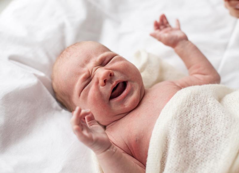 A newborn baby looks uncomfortable and cries while lying on a white bed.