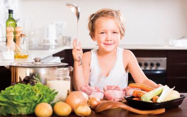Child nutrition for all ages and stages. little girl sitting at a counter full of vegetables holding a ladle and smiling