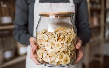person in apron holding large glass jar filled with banana chips