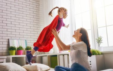 mom holding cape-wearing daughter up in the air