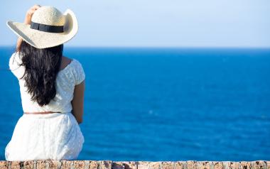 woman with long dark hair and a white sun hat sitting looking at the water