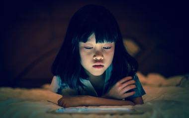 A young girl about 6 years old reads a tablet screen in an otherwise dark room