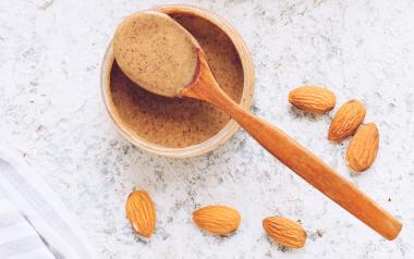A scoop of almond butter on a spoon poised over a small bowl surrounded by raw almonds.
