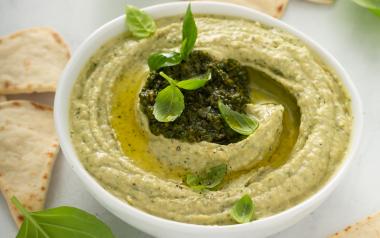 An image of creamy fava bean dip surrounded by bits of pita and some leaves of fresh basil.