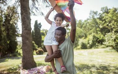 A dad with a child on his shoulders flying a kite in the summer sunshine.