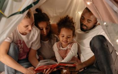 Parents and children spending time together in a makeshift indoor tent, reading a book