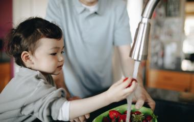 A child washes a strawberry under a kitchen faucet while a parent watches