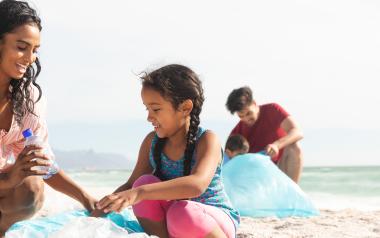 children and parents sit on a beach together with plastic bags for clean-up