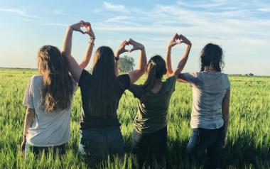 four teens face a field and link arms and hands to form heart shapes in the air