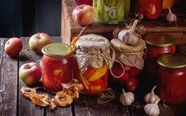 many homemade glass jars with preserved food (cucumbers, tomatoes, peppers), with garlic and fresh and dried apples over old wooden table