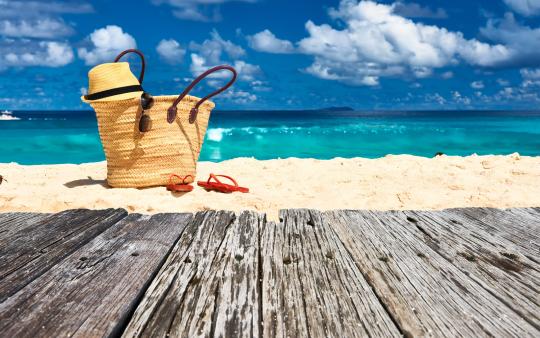 straw beach bag with hat on top sitting on the sand with water in distance and boardwalk in foreground