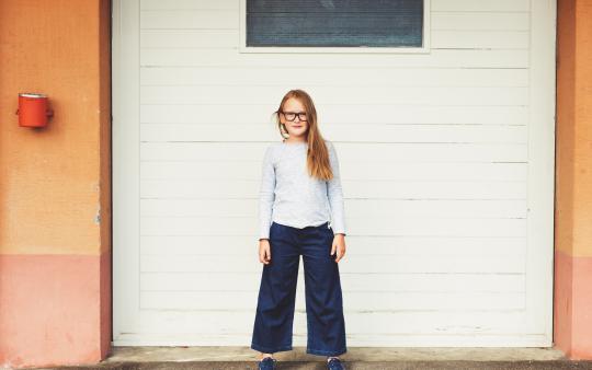 pre-adolescent girl standing in front of garage door wearing blue top, denim culottes and eyeglasses