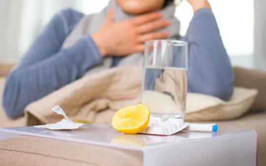 A woman holding her throat with a glass of water and lemon sitting on a table beside her