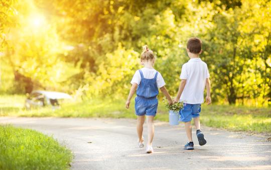 A brother and sister walking down a sidewalk each holding the handle of a bucket