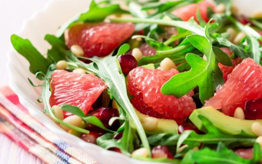 A white bowl containing arugula and grapefruit wedges, garnished with pine nuts