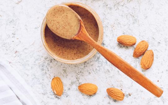 A scoop of almond butter on a spoon poised over a small bowl surrounded by raw almonds.