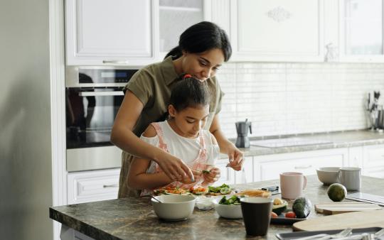 a mother standing behind her child, helping her chop vegetables