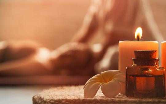 person being massaged in the background. a tray with a lit candle, bottle of oil and white flower in the foreground