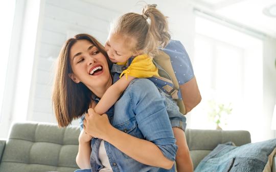 A woman on a couch is hugged by a little girl wearing a backpack