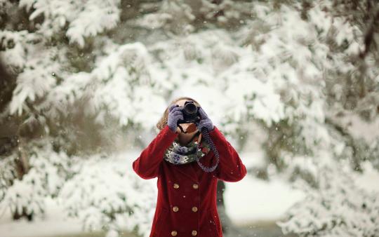 woman in red coat holds up a camera with pine trees covered in snow behind her