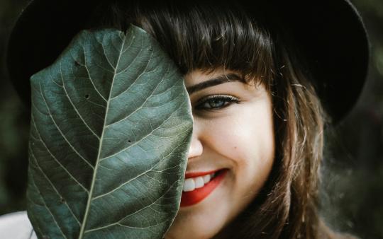 A woman wearing makeup smiles from behind a green leaf