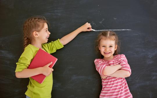 A girl marks the height of another child standing against a chalkboard