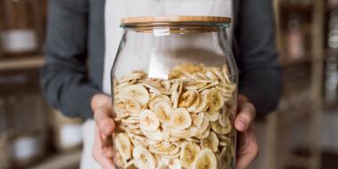 person in apron holding large glass jar filled with banana chips