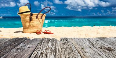 straw beach bag with hat on top sitting on the sand with water in distance and boardwalk in foreground