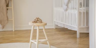 baby shoes placed on wooden stool standing in white kid room interior with crib, round rug 