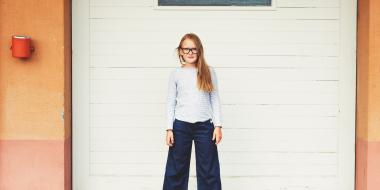 pre-adolescent girl standing in front of garage door wearing blue top, denim culottes and eyeglasses