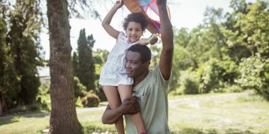 A dad with a child on his shoulders flying a kite in the summer sunshine.