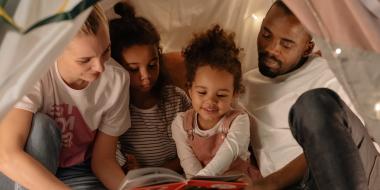 Parents and children spending time together in a makeshift indoor tent, reading a book