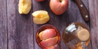 apples on a wooden table alongside a knife and jar of cider