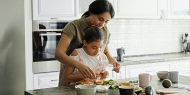 a mother standing behind her child, helping her chop vegetables